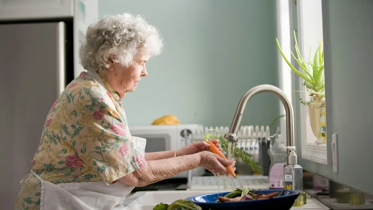 Oudere persoon drinkt medische drinkvoeding aan de keukentafel met hulp van een familielid